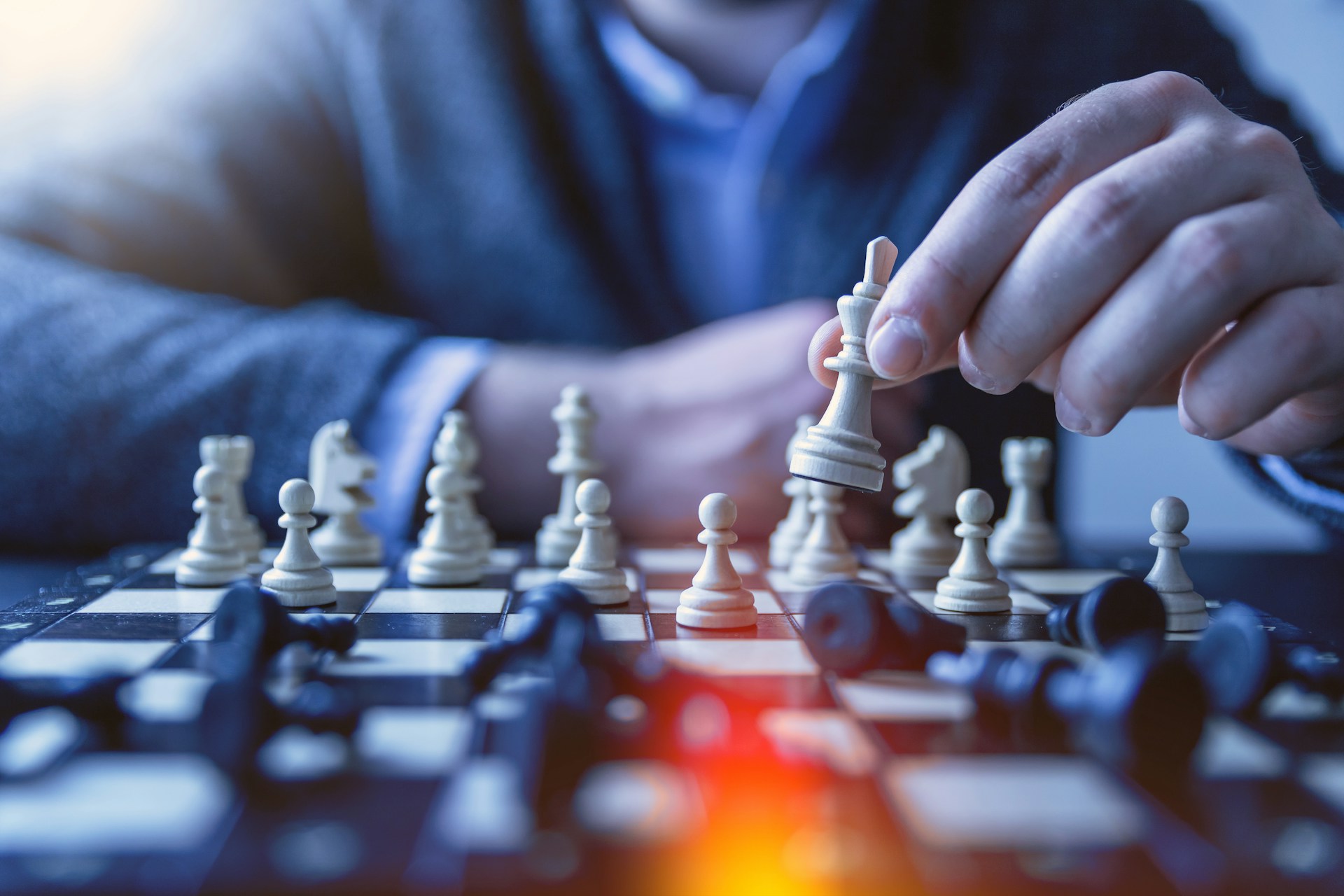 A hand lifts a white chess king on a board, with fallen black pieces blurred in the foreground.